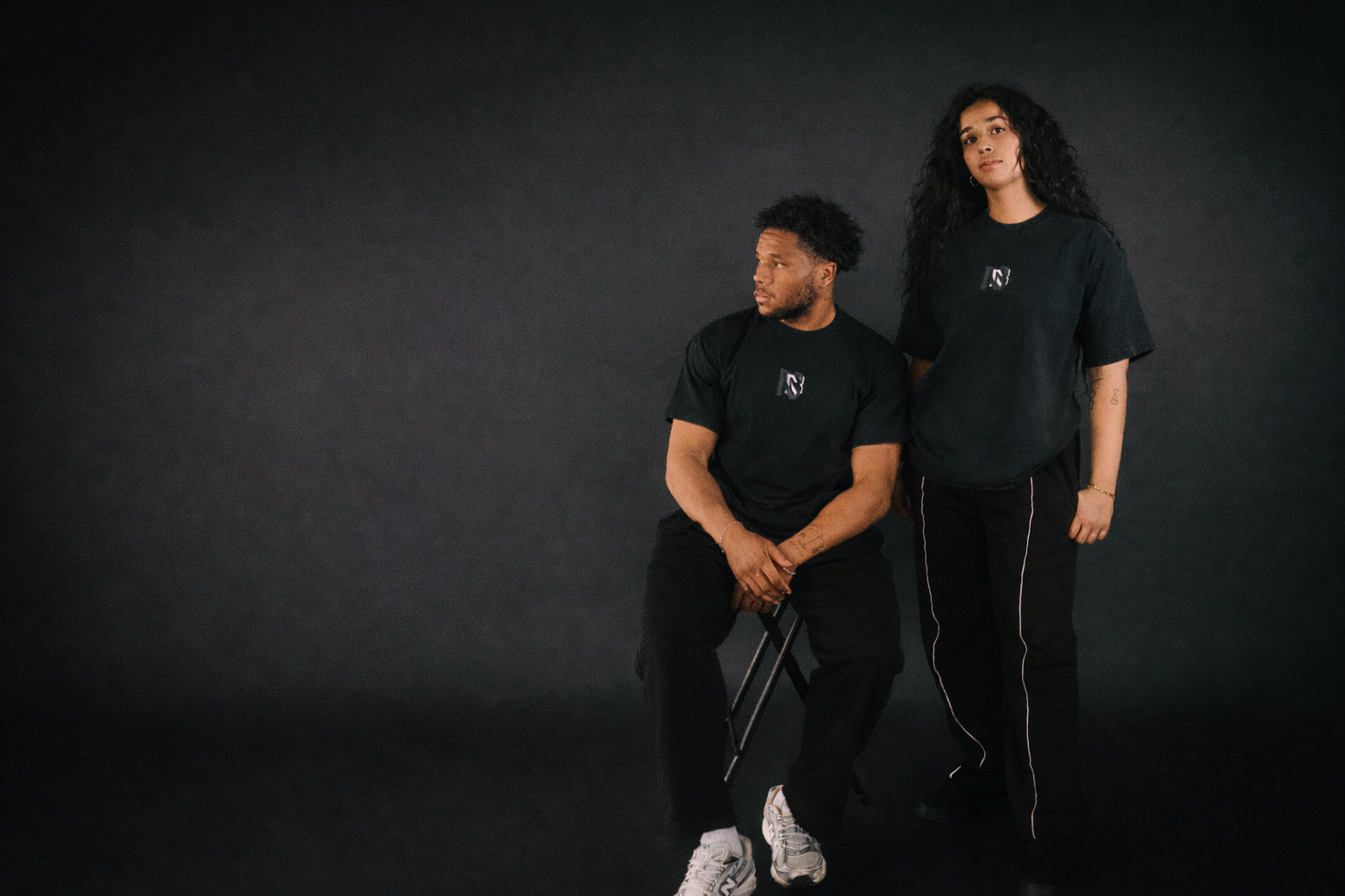 Two people wearing black t-shirts with a logo on a dark background