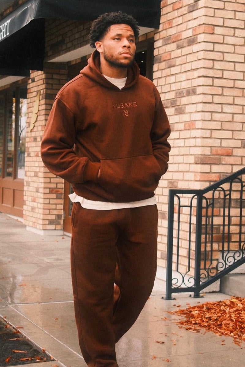 Man wearing a brown hoodie and pants standing in front of a brick building with 'Featherblade' sign.
