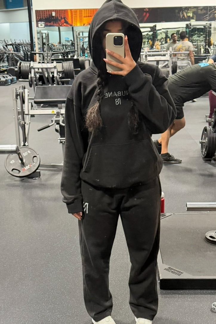Person taking a mirror selfie wearing a black hoodie and pants in a gym setting.